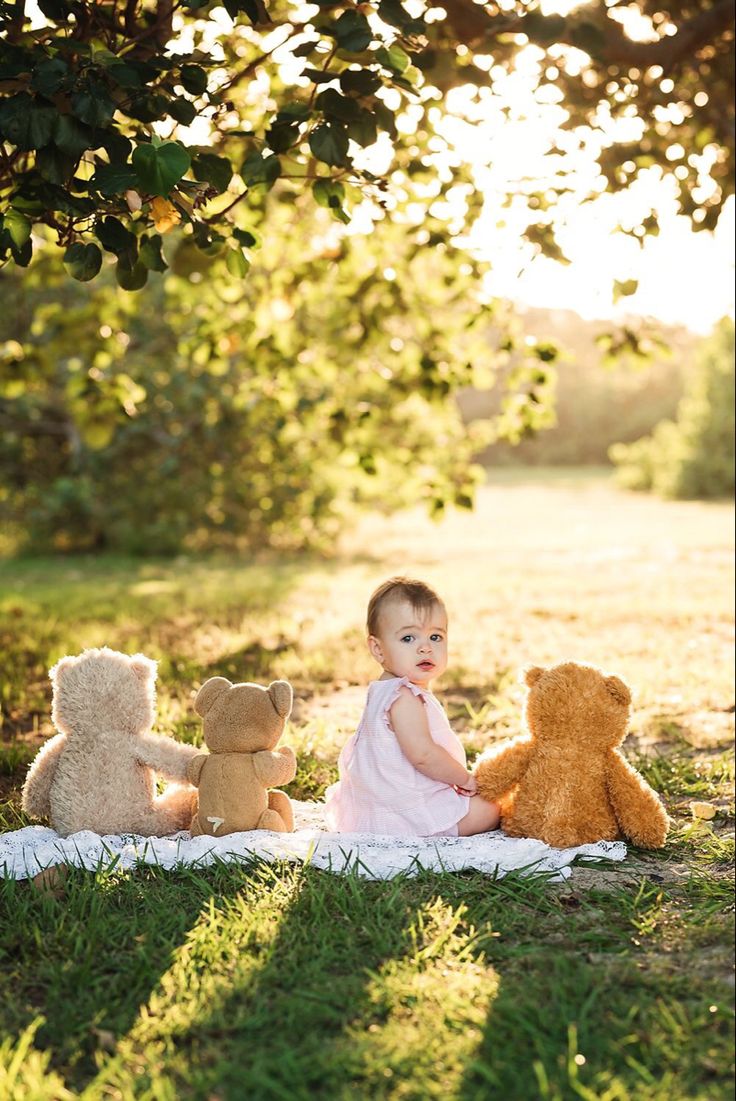 This may contain: a baby sitting in the grass with three teddy bears next to her on a blanket