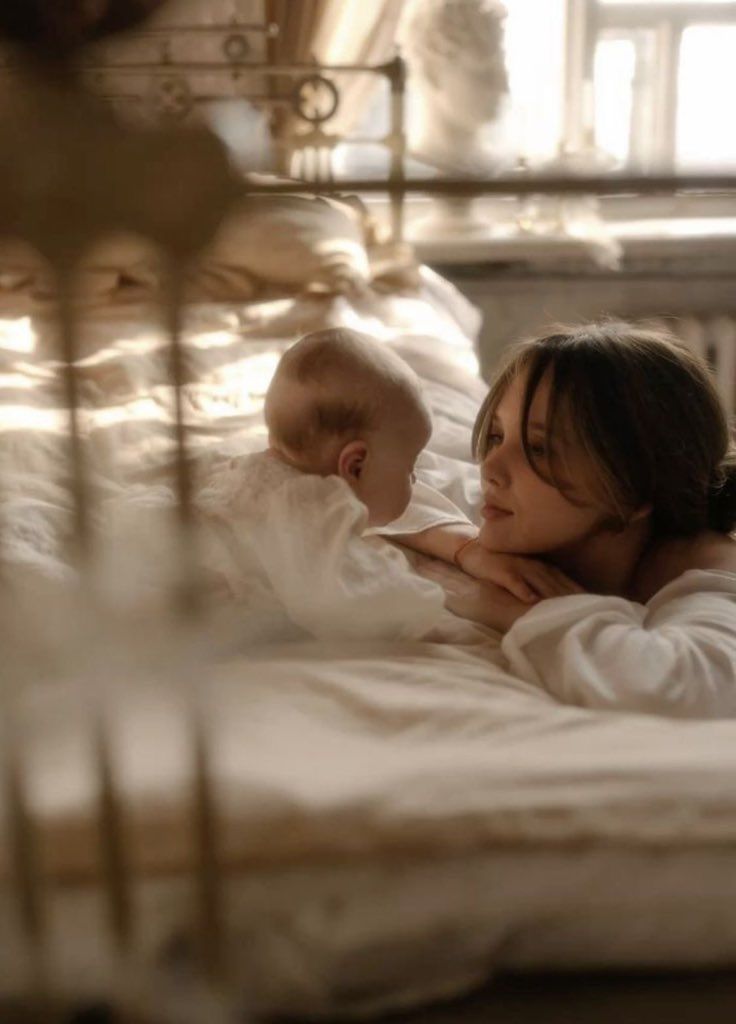 This may contain: a woman laying in bed with a baby on her stomach and looking at something off to the side